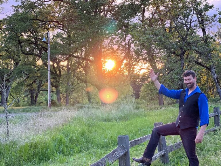 Dr. Jed Robinson, in a black vest, cowboy boots, and jeans,  offers a welcoming salutation from on a hiking trail at sunrise.