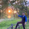 Dr. Jed Robinson, in a black vest, cowboy boots, and jeans, offers a welcoming salutation from on a hiking trail at sunrise.